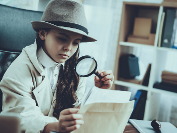 A young girl with a detective hat on looks at a paper through a magnifying glass