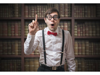 A person in a dress shirt, bowtie and suspenders, poses in front of bookshelves with a raised finger, as if realizing something