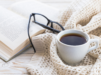 Warm knitted blanket with glasses, a cup of tea, and book on wooden table.