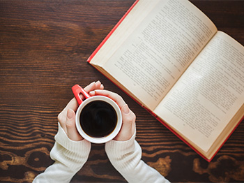 A table with a book and hands holding a mug.
