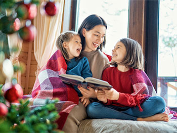 Adult reading a book to two children near a Christmas tree.