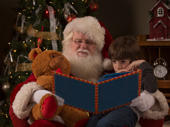 Santa Claus reading a story with a child on his lap with a teddy bear.