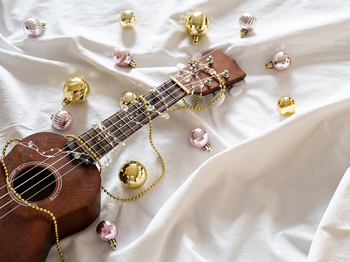 A ukulele lying on white sheets with shiney Christmas tree ornaments.