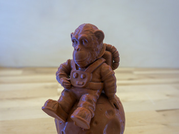 A 3D-printed monkey in a space costume sits on a wooden desk.