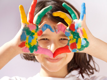 A child with paint-covered hands making a heart shape with their hands in front of their face.