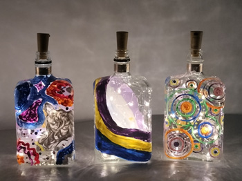 Three corked glass bottles with decorative paint on the outside and a string of lights inside.