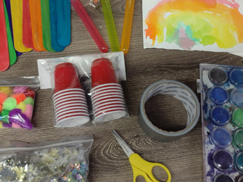 An assortment of crafting materials like popsicle sticks, scissors, and paints rest on a wooden table top.
