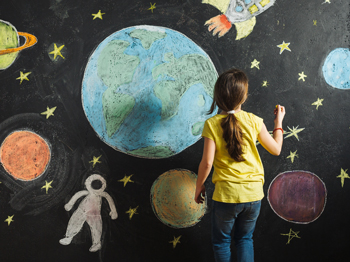 A child stands in front of a colourful chalkboard with illustrated depictions of plants, stars, spaceships, and an astronaut.