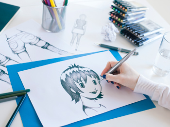 A person is drawing a manga character on paper with art supplies nearby on a table.