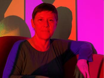 A photograph of artist Lyla Rye sitting in front of a multi-colour projection