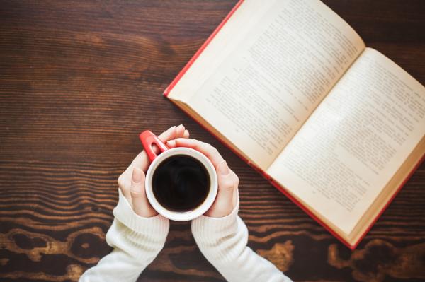 Hands holding a mug of coffee and open book to the side.