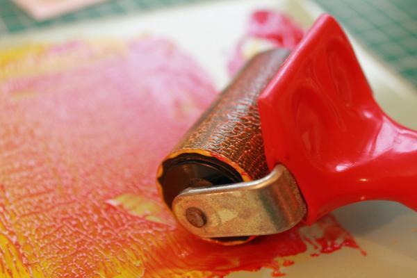 A brayer rolls out red ink on a yellow piece of paper.
